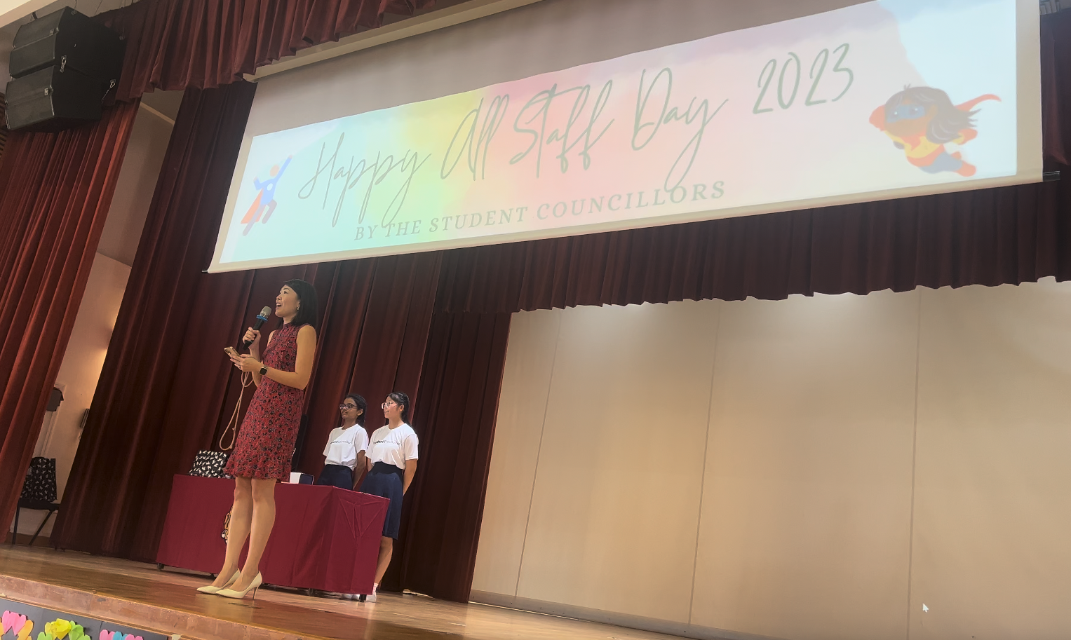 A woman speaks into a microphone on stage with two students behind her under a Happy All Staff Day 2023 banner.
